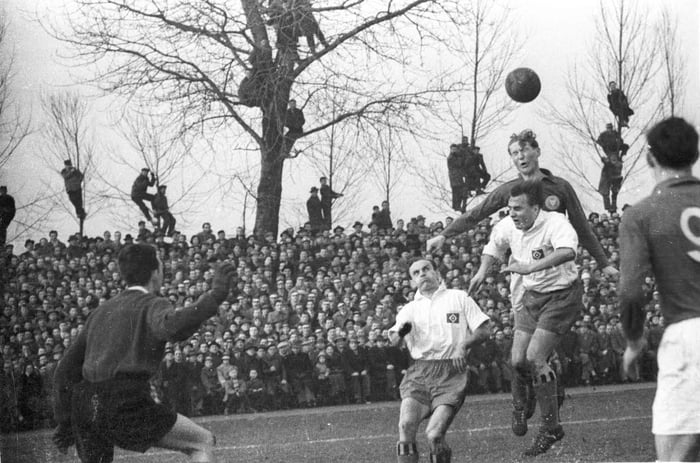 Two German clubs playing football as fans look over on trees in 1957
